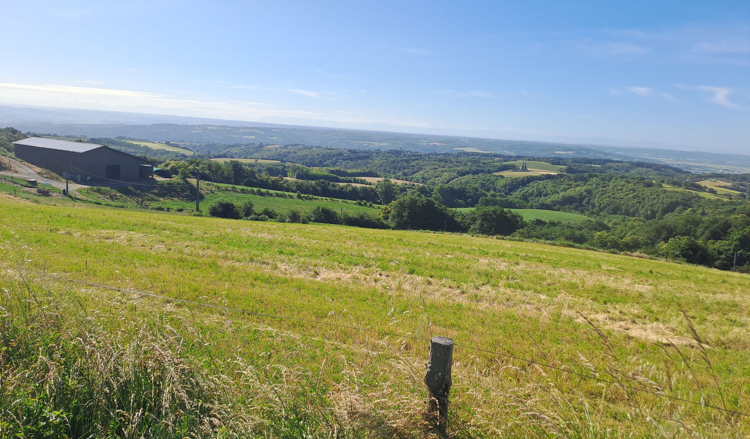 Vue panoramique d'une campagne vallonnée sous un ciel bleu clair, avec un champ d'herbe verte au premier plan et une grange sombre sur la gauche. Saint-Romain-en-Gal - Rhône. France