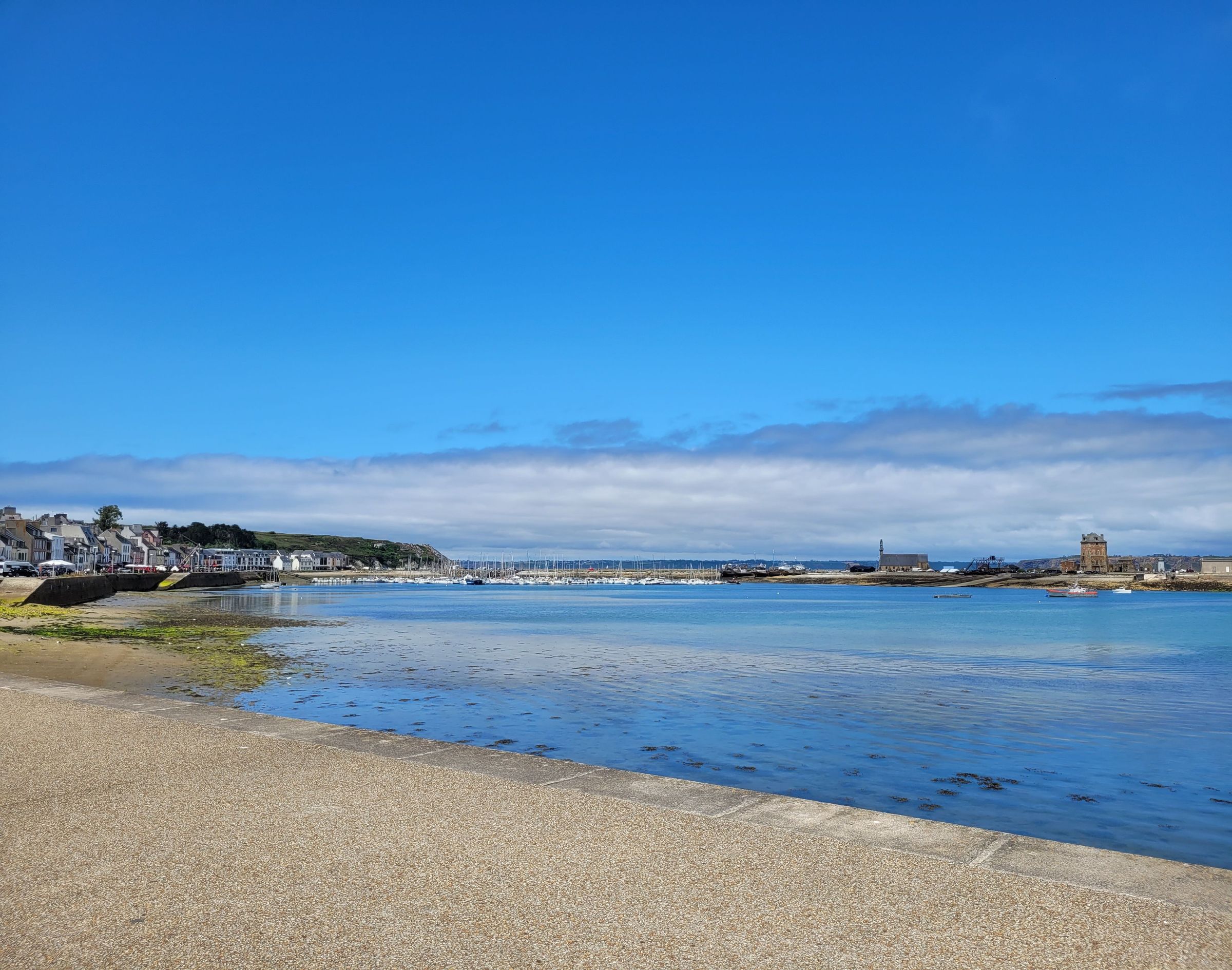 Vue panoramique d'un port de pêche breton avec des maisons blanches, des bateaux amarrés et un phare au loin, sous un ciel bleu clair avec quelques nuages blancs.